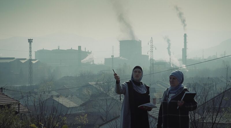 Two women stand outside in a city, while large pipes billow smoke in the background.