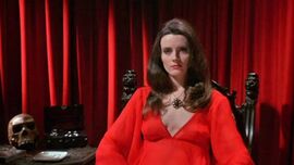 Woman with dark hair and a red dress sits on a chair in front of a red curtain. A skull and box sits on a table next to her