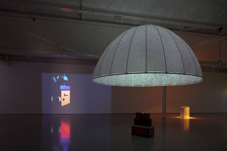 From Hiraki Sawa's exhibition. A blue, dome-like structure which has been separated by wire into segments, hangs from the ceiling. In the background, a screen shows a picture of an orange camera.