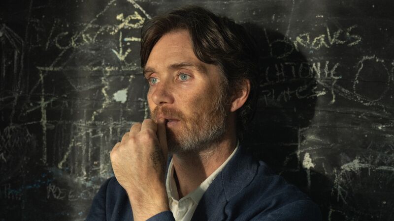 A man with a beard leans against a blackboard, looking pensive.