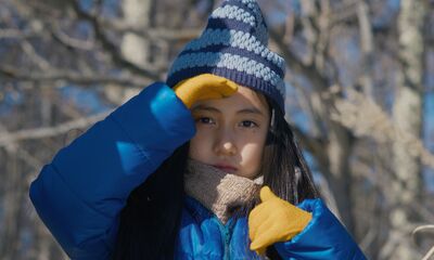 A young girl standing outside with yellow gloves and a blue jacket on shields her eyes from the sun.