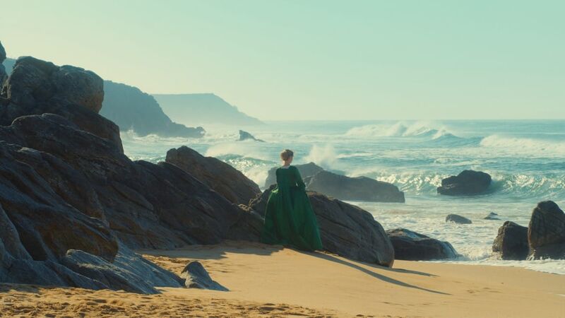 A woman in historical dress watches waves on a beach.