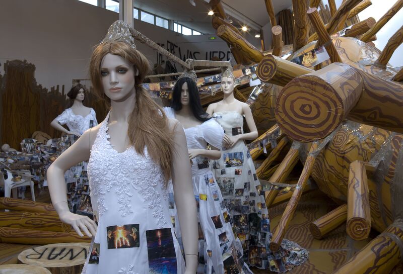 From Thomas Hirschhorn's exhibition. Mannequins are dressed in wedding dresses which are covered in pictures of burning buildings. There is a large, fake, chopped down tree on the ground behind them.