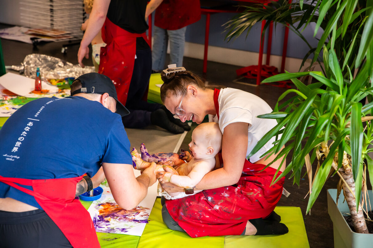 Parents wearing red aprons and holding a baby who is covered in purple paint
