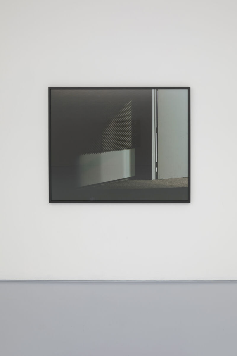 A photograph in a slim black frame is hung on a white wall. We can see in the photograph the bottom of a doorframe, with the door open and a small chink of light coming into the room. 