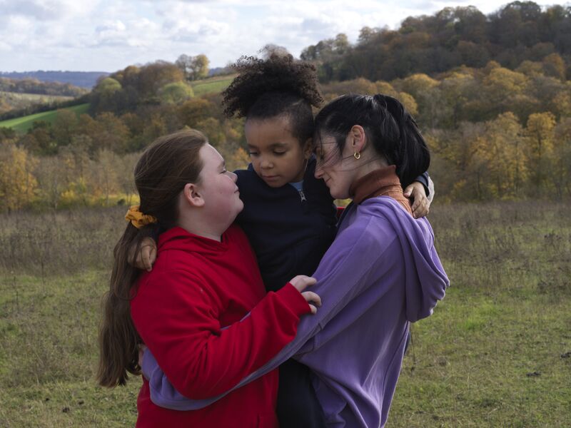 A woman hugs two little girls in a field.