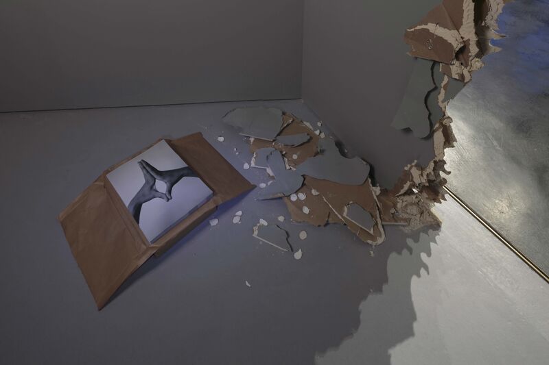 From Margaret Salmon's exhibition, Hole. A wall has been smashed, leaving debris on the ground. Next to the debris, there is a photograph of two hands.