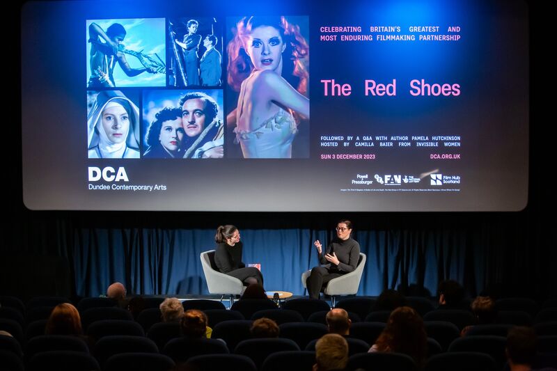 Two women in black sit on a stage at the front of DCA cinema, discussing a film. The screen behind them promotes a screening of The Red Shoes.