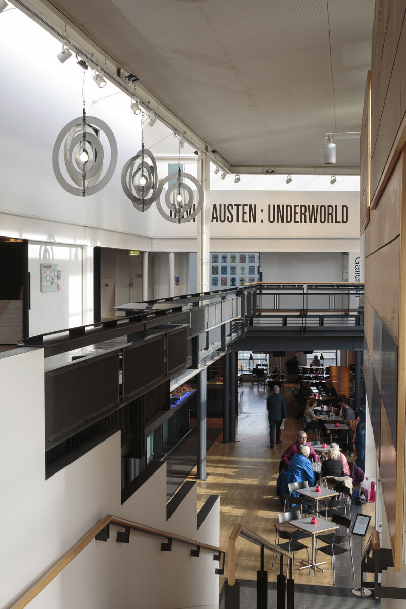 Image shows the interior of DCA's foyer, facing towards the gallery entrance from the top of the stairs. We can see the title of the exhibition above the entrance to the gallery, and on the left hang three suspended artworks which are white and consist of concentric flat rings. Down the stairs we can see visitors to the cafe bar sitting at chairs and tables. 