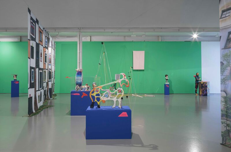 An image from Shonky: The Aesthetics of Awkwardness shows a colourful statue of a ram sitting on a blue box. In the background, there is a picture of a building printed on fabric. The wall at the end of the room is green.