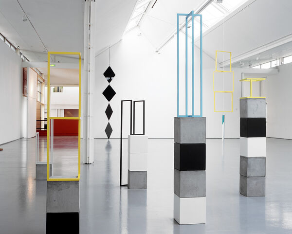 Sculptures from Camilla Løw's exhibition. Black cubes are stacked on top of each other, with yellow and blue coloured rectangular frames placed on top of them. 