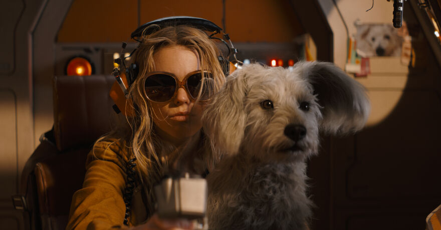 A person wearing a headphones sits inside a spacecraft cockpit beside a small white dog, surrounded by illuminated controls and warm interior lighting.