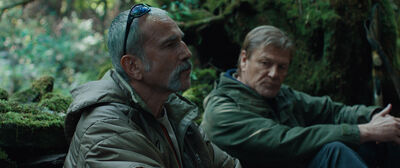 Two men sit outside in a forest, wearing khaki jackets
