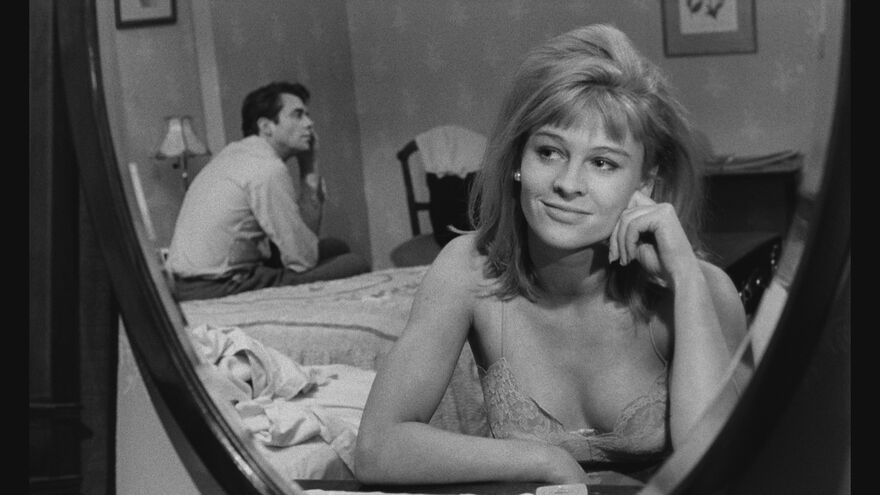 A blonde woman with a fringe smiles while sitting on one side of a bed while a man sits on the other side.
