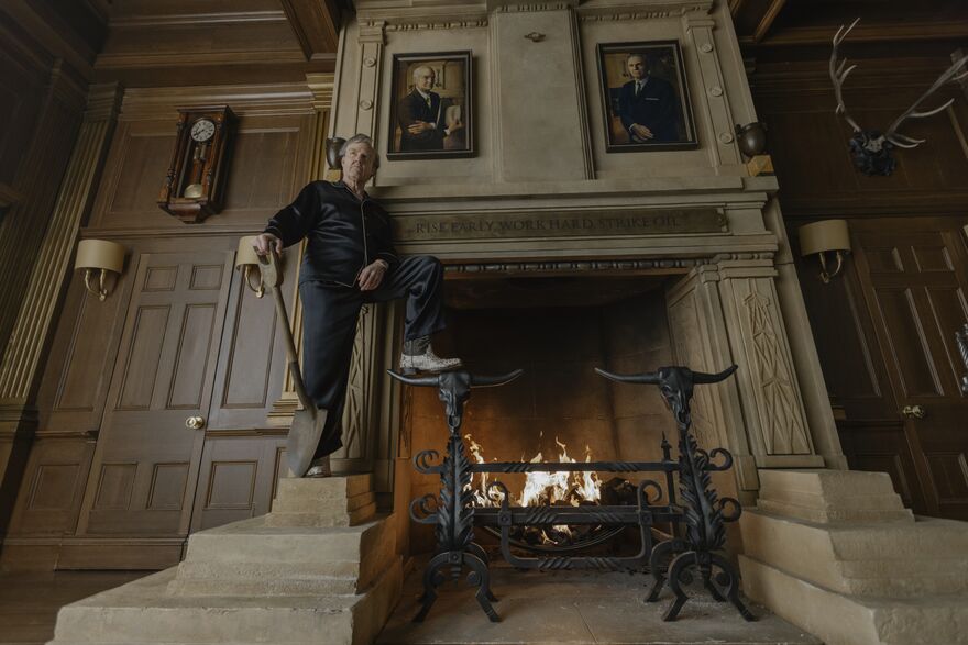 An old man stands next to a fireplace holding a shovel