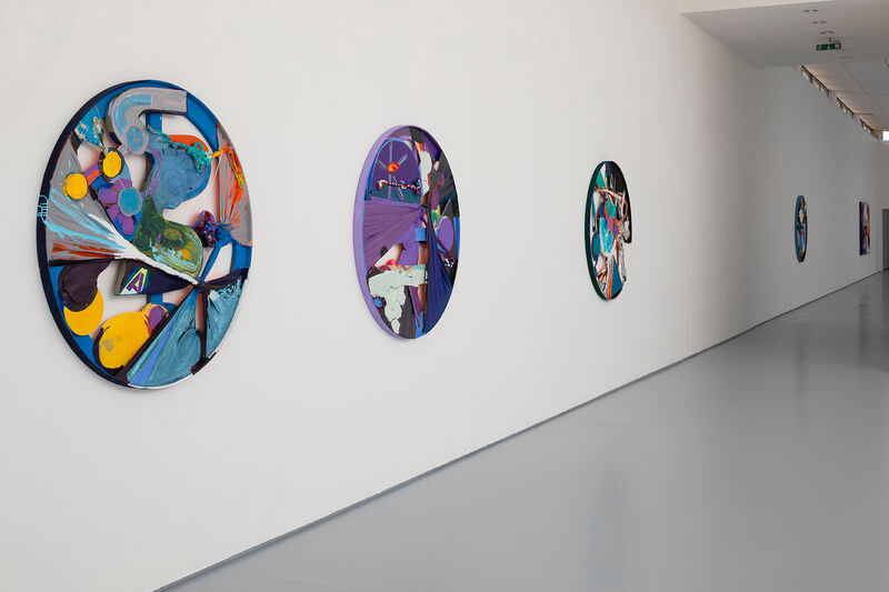 Installation photograph showing a series of round works on a white wall, going into the distance. 