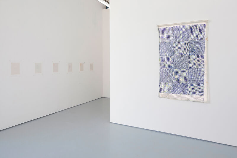 An installation photograph of artworks on paper. A line of a4 sheets on a white wall can be seen to the left of the image. To the right, a mainly blue artwork with a check-like pattern hangs on its own. 