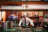 A young man wearing a white shirt and black tie leans on the bar of a traditional bar with 90s images propped against the back 