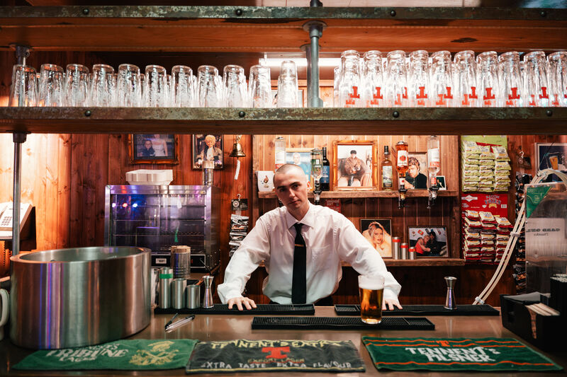 A young man wearing a white shirt and black tie leans on the bar of a traditional bar with 90s images propped against the back 