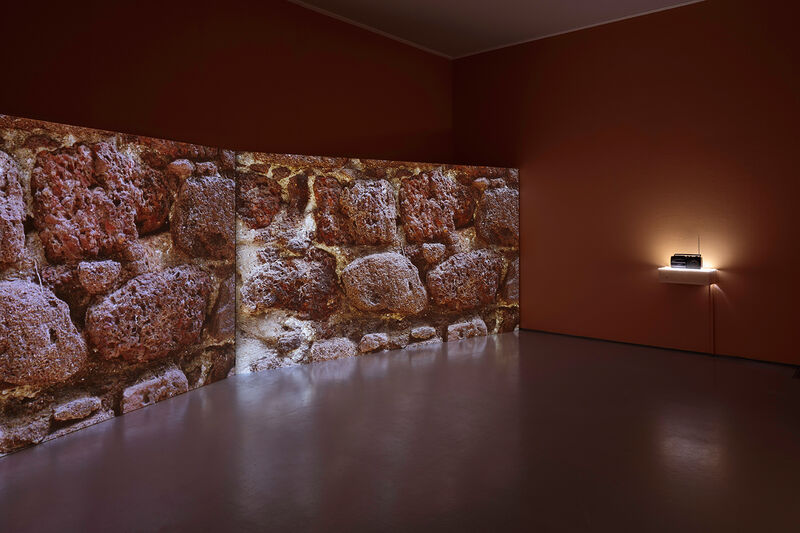 A photograph of a two channel video installation, with two screens leaned on the floor against a dark orange wall. On the screens can be seen a close up of some rough red rocks. To the left of the image, a black 1980s style radio sits on a white shelf at waist level. 