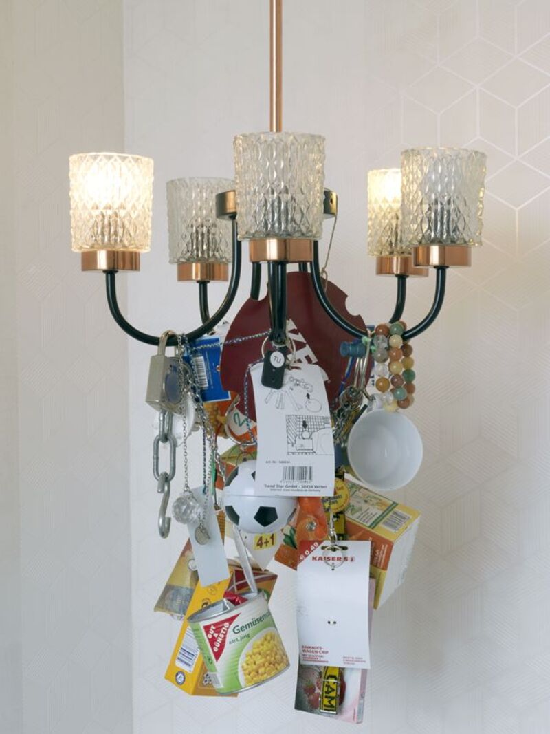 A 5-way ceiling light from Manfred Pernice's exhibition is decorated with mundane objects -  a can of sweetcorn, a padlock, bangles, a mug, a keyring, a KFC lid.