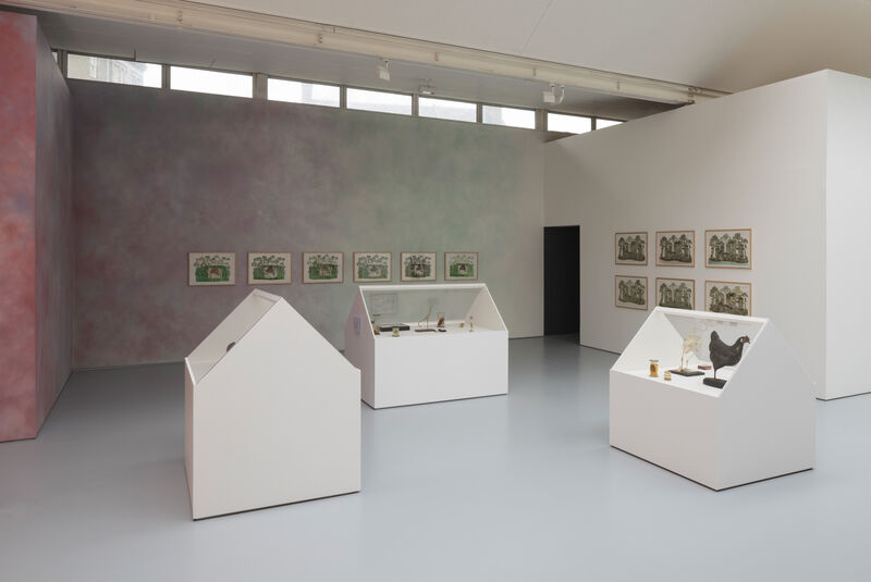 Three plinths with glass apexes can be seen in the centre of the photograph, displaying small objects as though a museum display. Behind can be seen a wall painted with mottled pink and blue, with a row of six paintings. To the right there is a small dark doorway leading to a video booth. On the far right wall can be seen a grid of six paintings. 