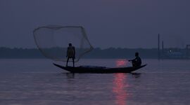Two figures on a fishing boat at sunrise, a fishing net thrown in the air.