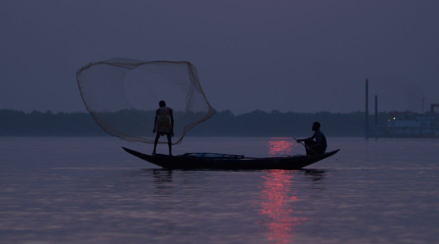 Two figures on a fishing boat at sunrise, a fishing net thrown in the air.
