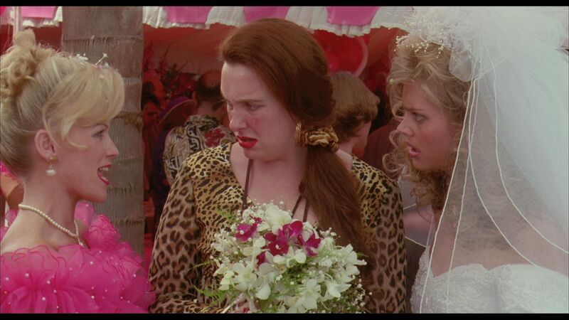  A young woman holding a bouquet looks in disgust at a woman in a pink fluffy outfit, while a bride looks at the first young woman in disgust.