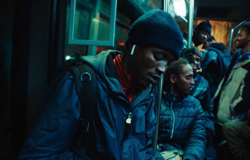 A man wearing a rain jacket, a hat and airpods sleeps standing up on a bus in the dark
