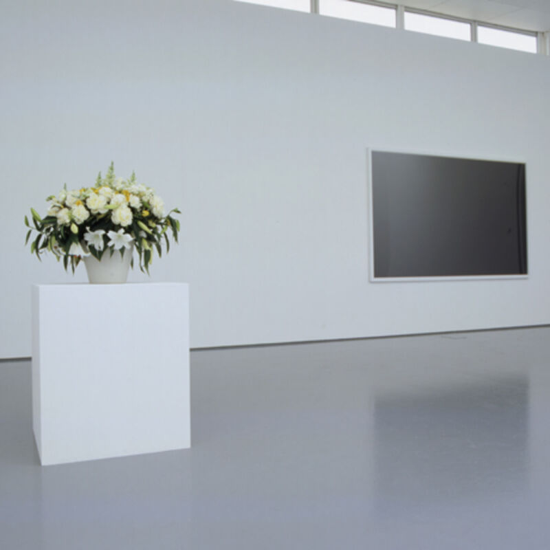 An image from Marine Hugonnier's shows a white pot which is filled with a large display of drooping white and yellow lilies and roses. In the background, there is a large, black board on the wall.