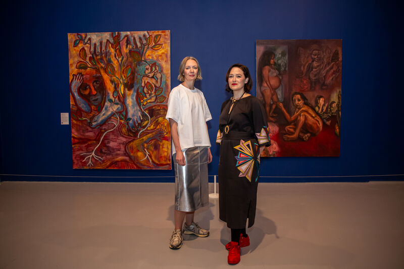 Two white women look directly at the camera a blue wall with large images paintings of muted colours in the backgroud