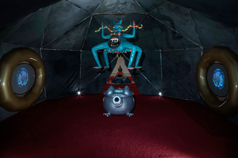This photograph is taken inside a geodesic dome containing video and ceramic elements. A ceramic figure at the centre of the photograph has four legs, two arms, a gaping mouth in it's belly, three eyes and is bright blue in colour. 