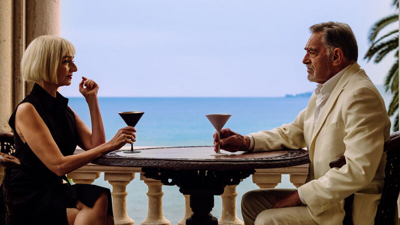Woman and man sitting across from each other at a table, each holding a cocktail glass. There is a view of the sea in the background
