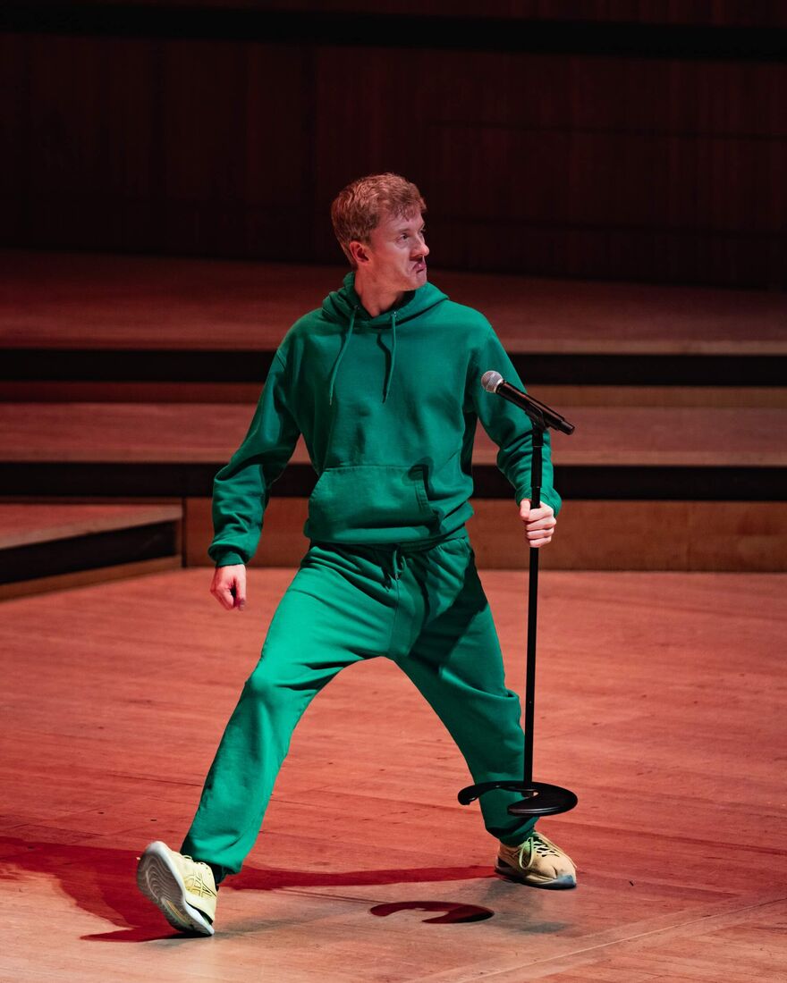 James Acaster performs comedy on stage while wearing a green tracksuit