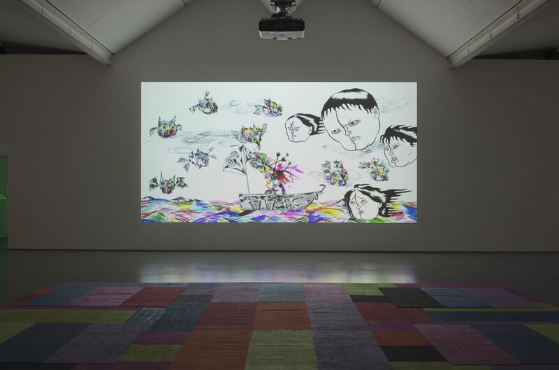 A projection from Hideyuki Katsumata's exhibition. The image shows floating heads against a giant multi-coloured sea. There is a checkered, rainbow coloured rug on the floor of DCA Galleries.