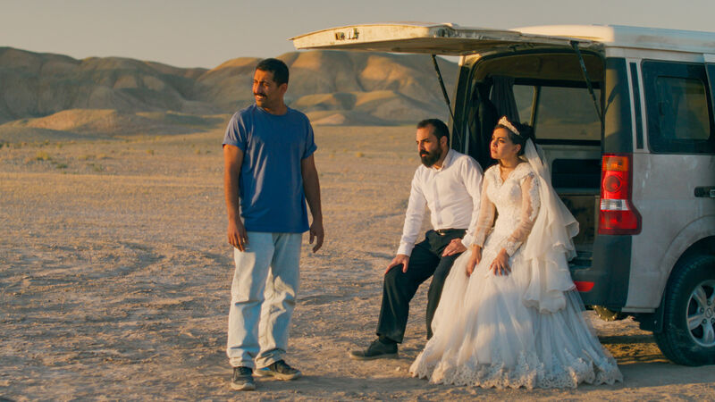 Three people stand in a desert with a landcover car with them. This is two men, and one woman in a wedding dress