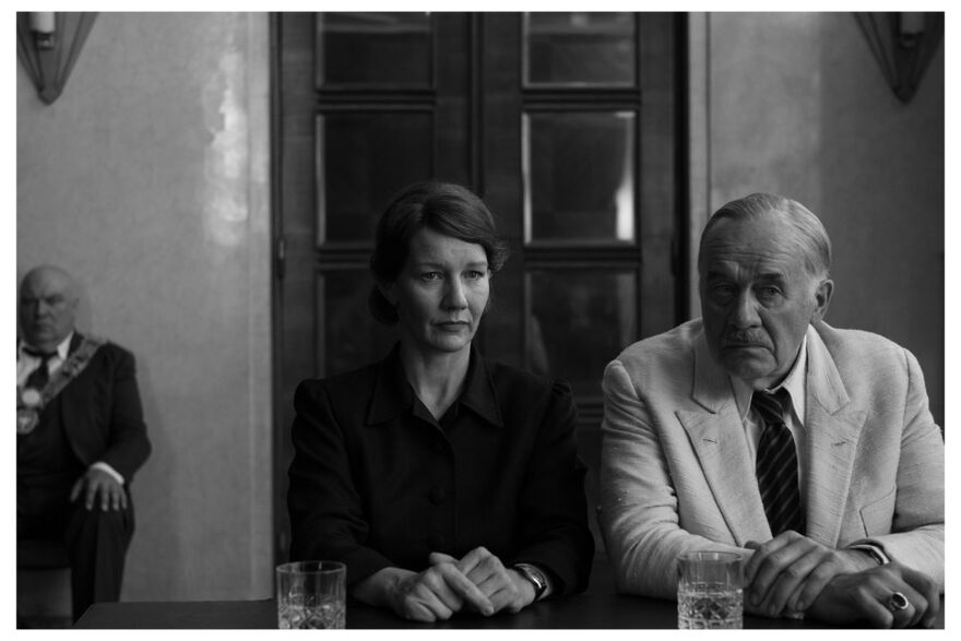 A woman and man sit facing towards us while another man sits further behind watching them