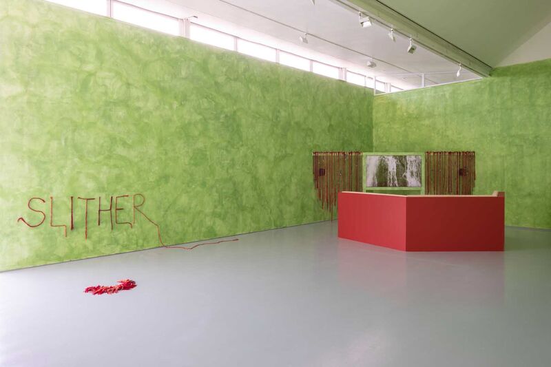 A gallery with green walls. In the corner is a video screen, with a red seating area in front of it. The word 'Slither' appears on the wall.