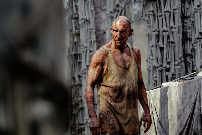 A bald man, covered in blood and wearing a dirty vest, stands next to a statue made of bones.