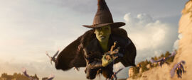A green-skinned woman flies on a broomstick.