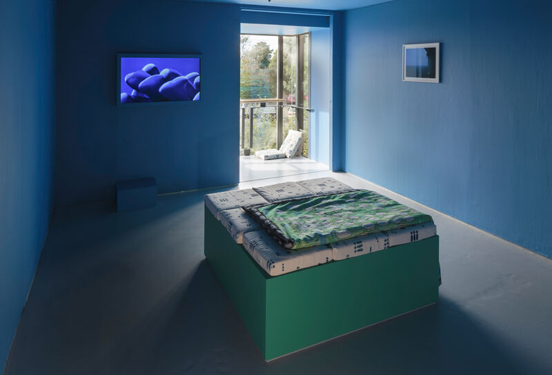 Photograph of the auxiliary gallery at the back of gallery two. A seat with cushions and throws is the the middle of a blue painted room, the plinth painted green. On the right hand wall can be seen a framed photograph. Straight ahead on the left is a monitor on the wall showing a blue image. The window is bright and there are cushions on the floor next to it. 