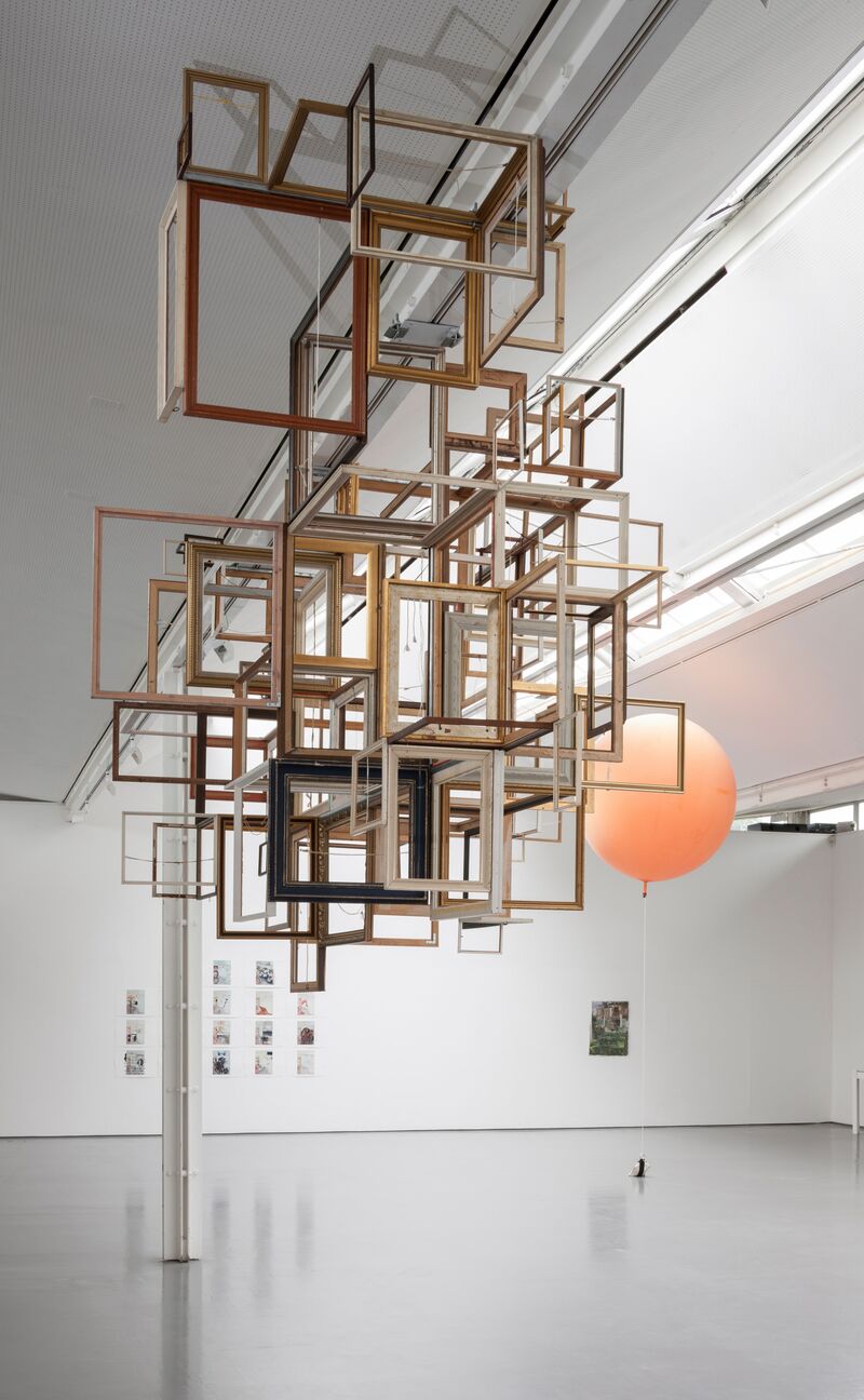 An installation from Continue Without Losing Consciousness shows many portrait frames stuck together to make a 3D structure, which is suspended from the ceiling. In the background, a large orange balloon can be seen.