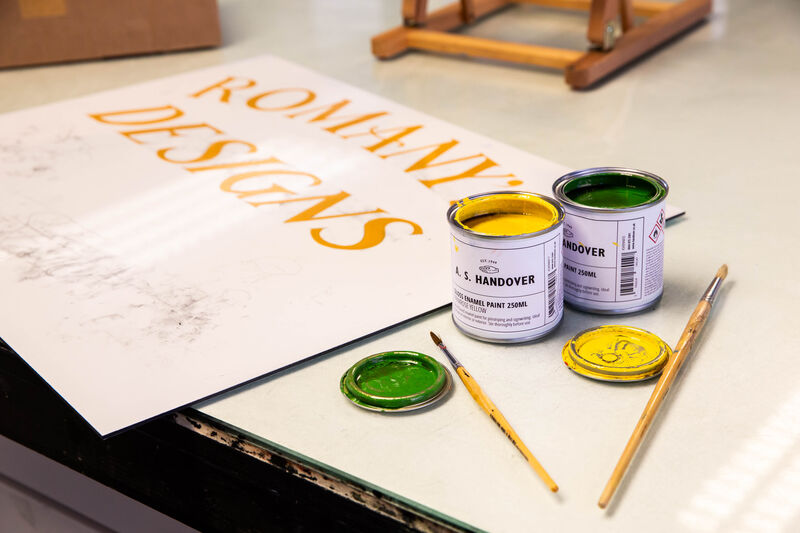 Sign painted with gold text with open pots of paints and brushes beside it