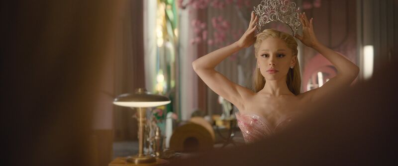 A woman with blonde hair wearing a pink dress places a diamond-studded crown on her head.