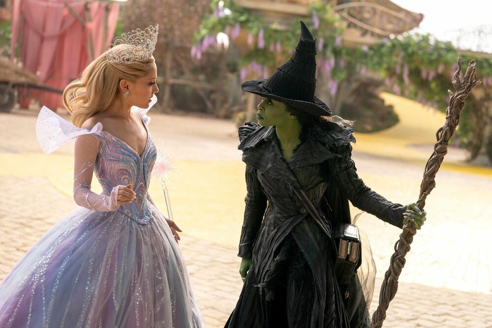 A woman with a wand, a tiara and a purple dress stands next to a woman wearing a witches hat who has green skin.