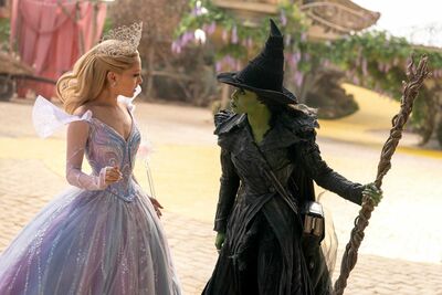 A woman with a wand, a tiara and a purple dress stands next to a woman wearing a witches hat who has green skin.
