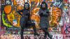 Two figures dressed in black, wearing gorilla masks, stand in front of a wall covered in graffiti. One has their arms folded and the other an arm on their hip.  