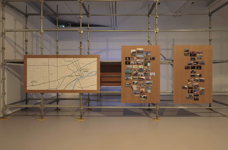 Three boards suspended on scaffolding as part of Mike Kelley's exhibition. One shows a map. The other two are a collection of postcards.