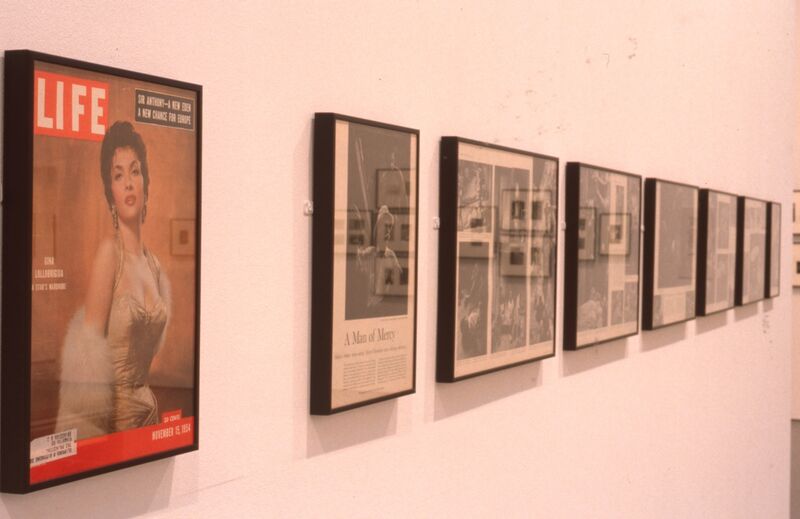 Framed newspaper and magazine clippings hang on a white wall, including a 'Life' magazine cover with a portrait of a lady on the front.